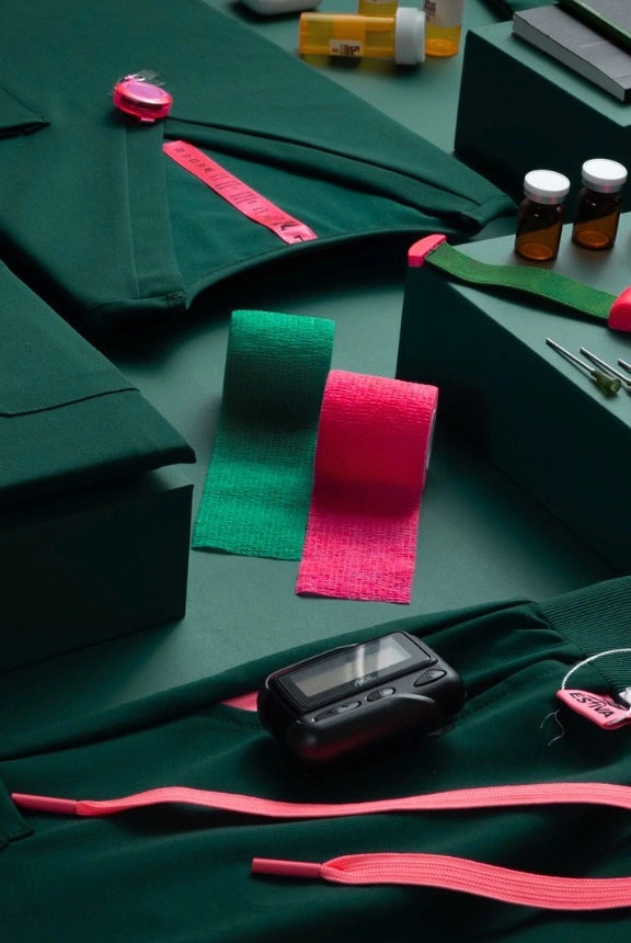 Dark green Estiva scrubs with pink drawstring, pink measuring tape, and bright pink and green medical wraps in a flat lay setup