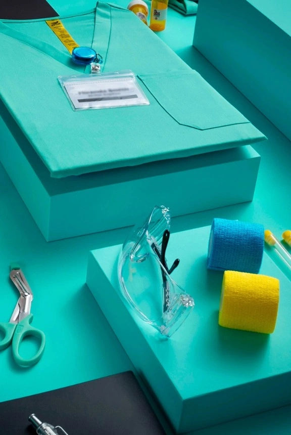 Flat lay of teal Estiva scrub top with medical accessories and yellow drawstring